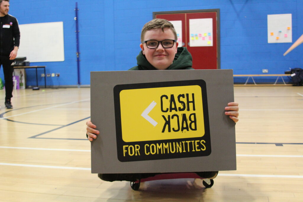 Young person wearing glasses smiling. Holding a CashBack for Communities Sign.