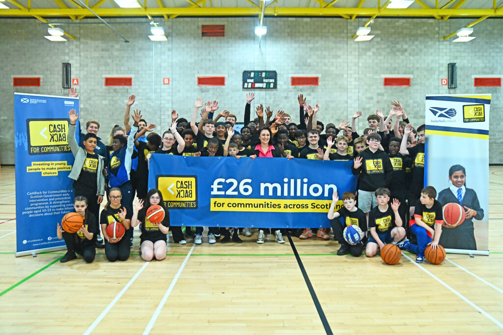 Minister Siobhian Brown with young people inside sports hall holding banner with CashBack for Communities logo reading '£26 million for communities across Scotland'